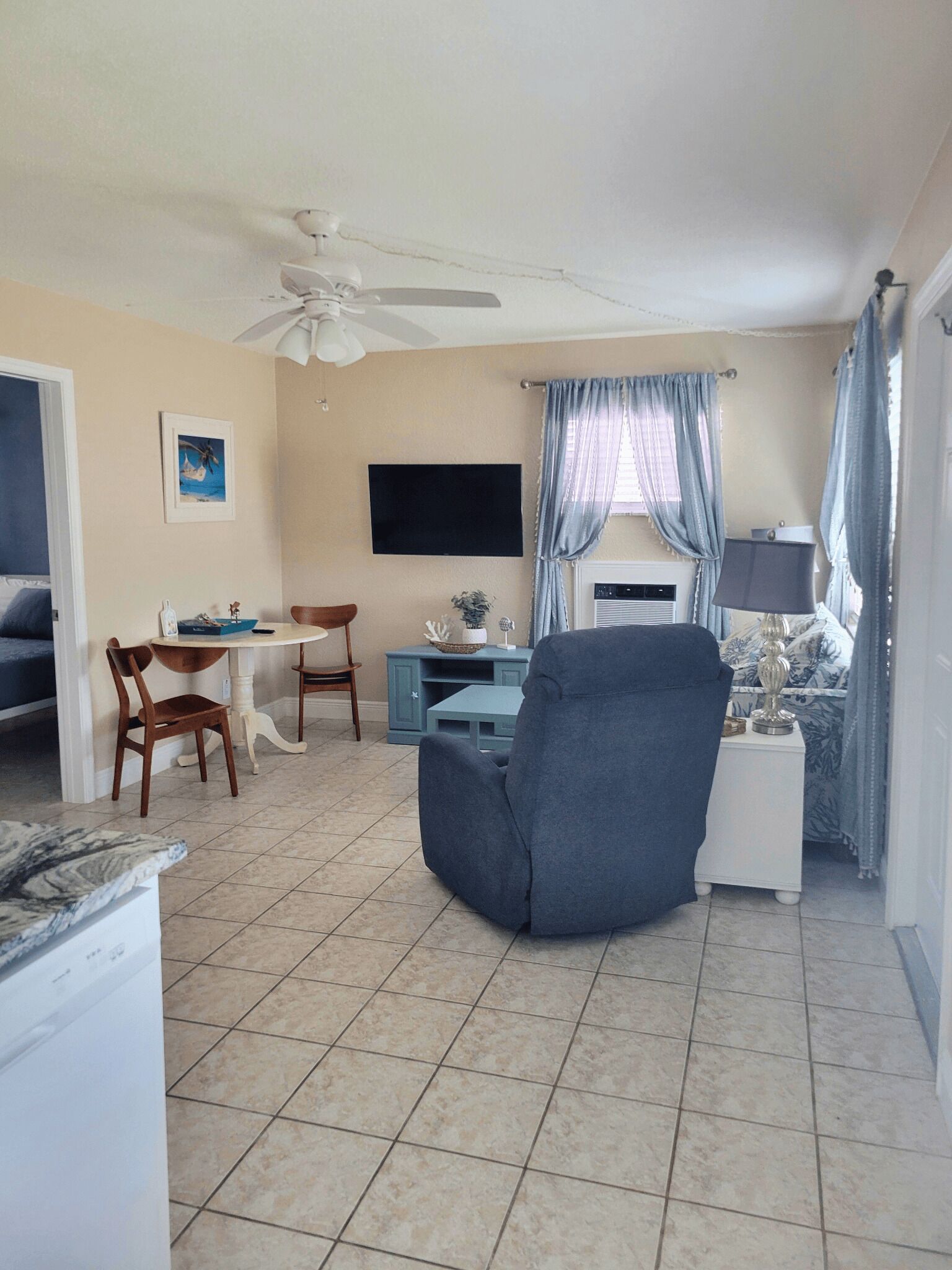 A living room with a ceiling fan, a TV, a dining table with chairs, a blue cabinet, and a window with curtains.