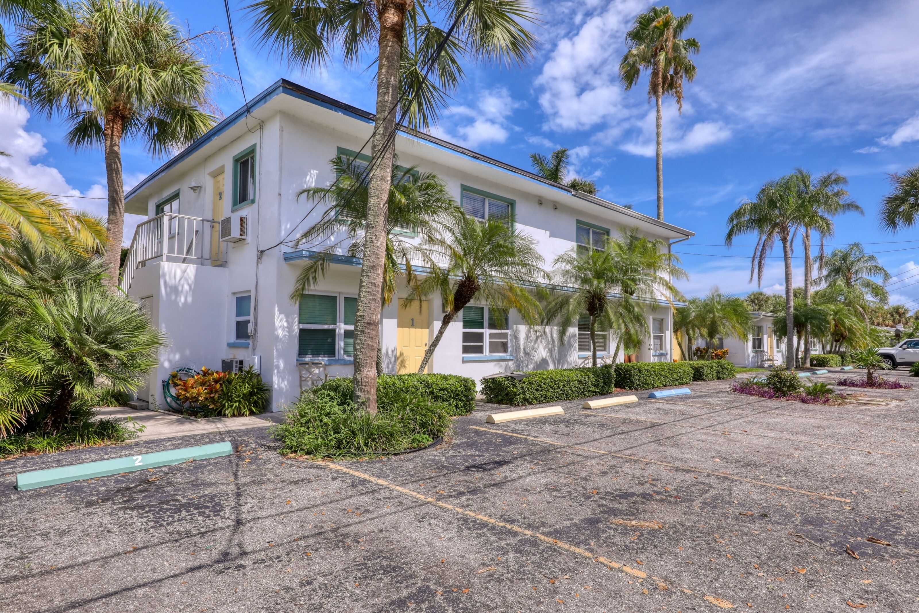 A white building with a balcony, surrounded by palm trees and a parking lot.