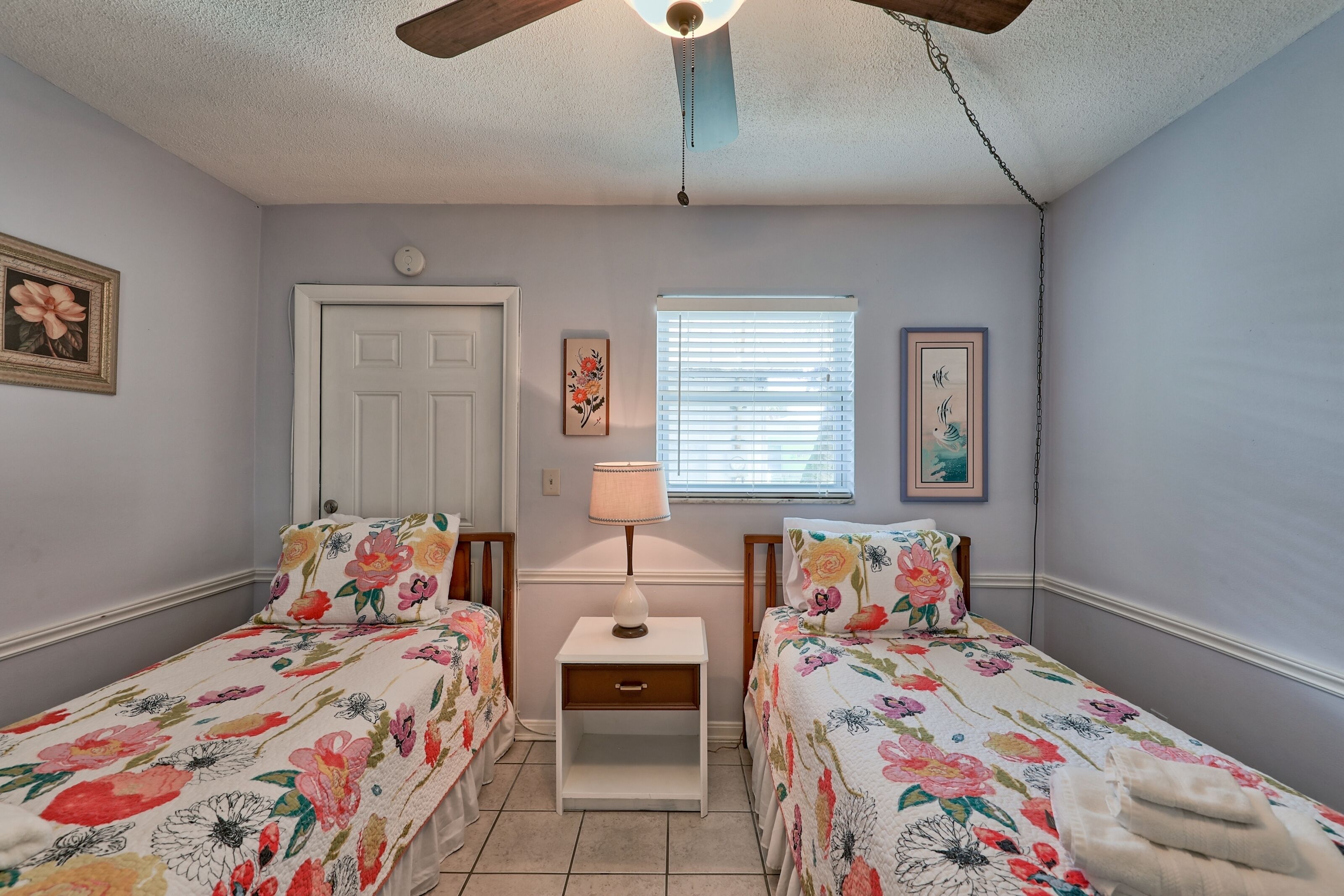 A bedroom with two beds, a nightstand, a ceiling fan, a window with blinds, and a door.