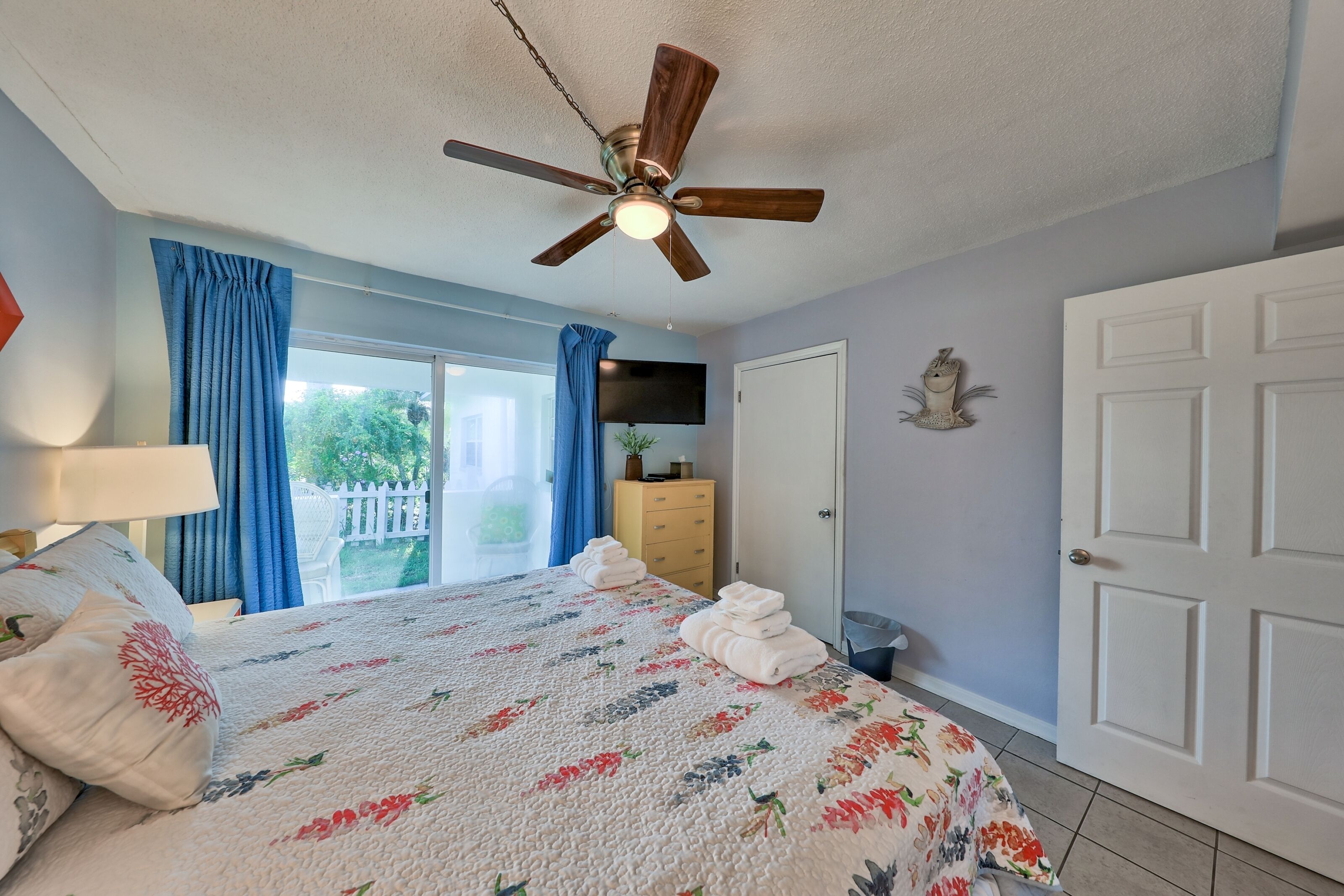 A bedroom with a bed, a ceiling fan, a TV, a dresser, and a door.