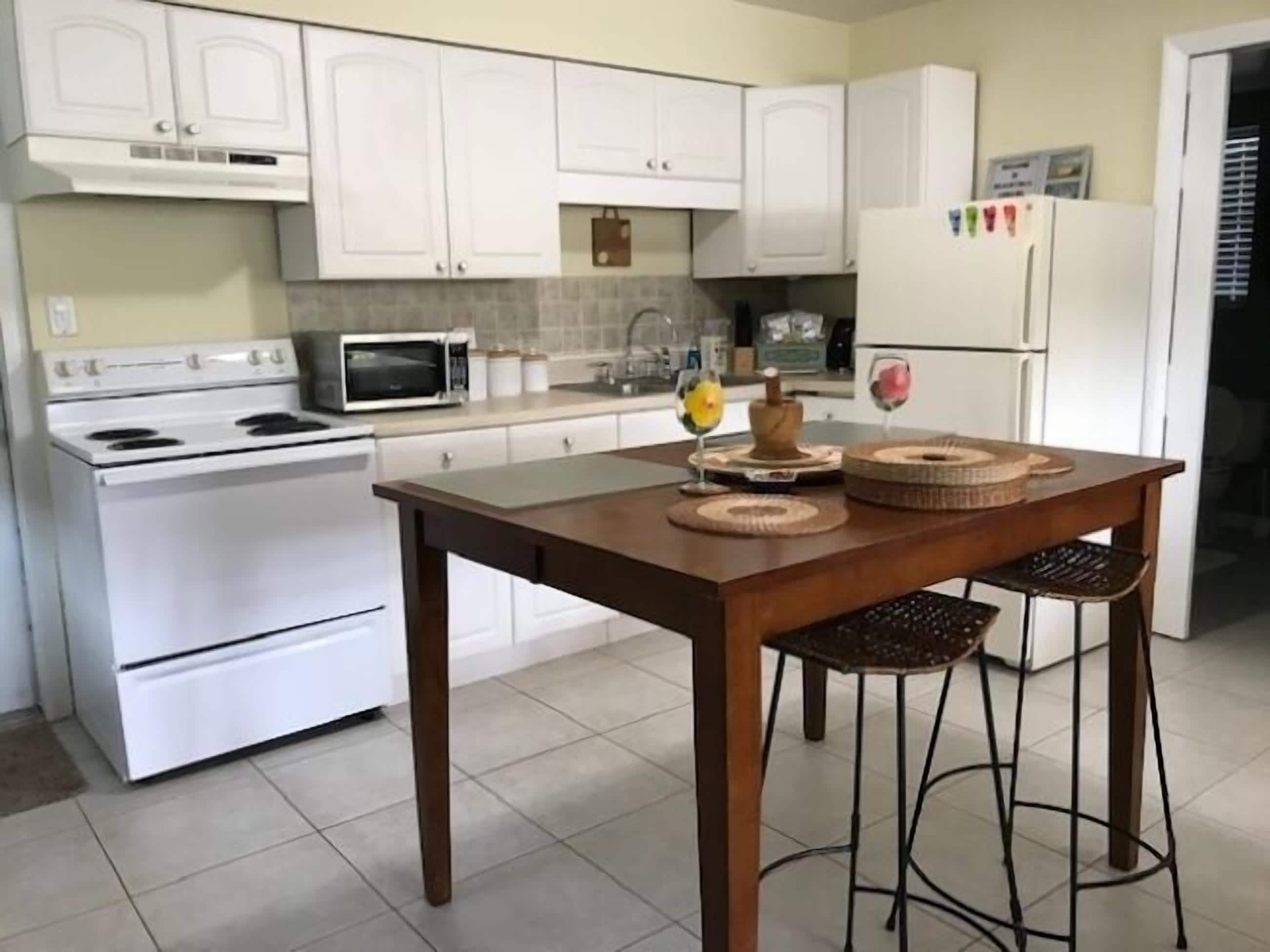 A kitchen with white cabinets, a stove, a microwave, and a refrigerator. There is a wooden table with two chairs and decorative items on it.