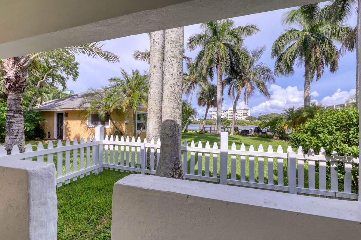 A white picket fence, palm trees, and a building with a yellow facade.