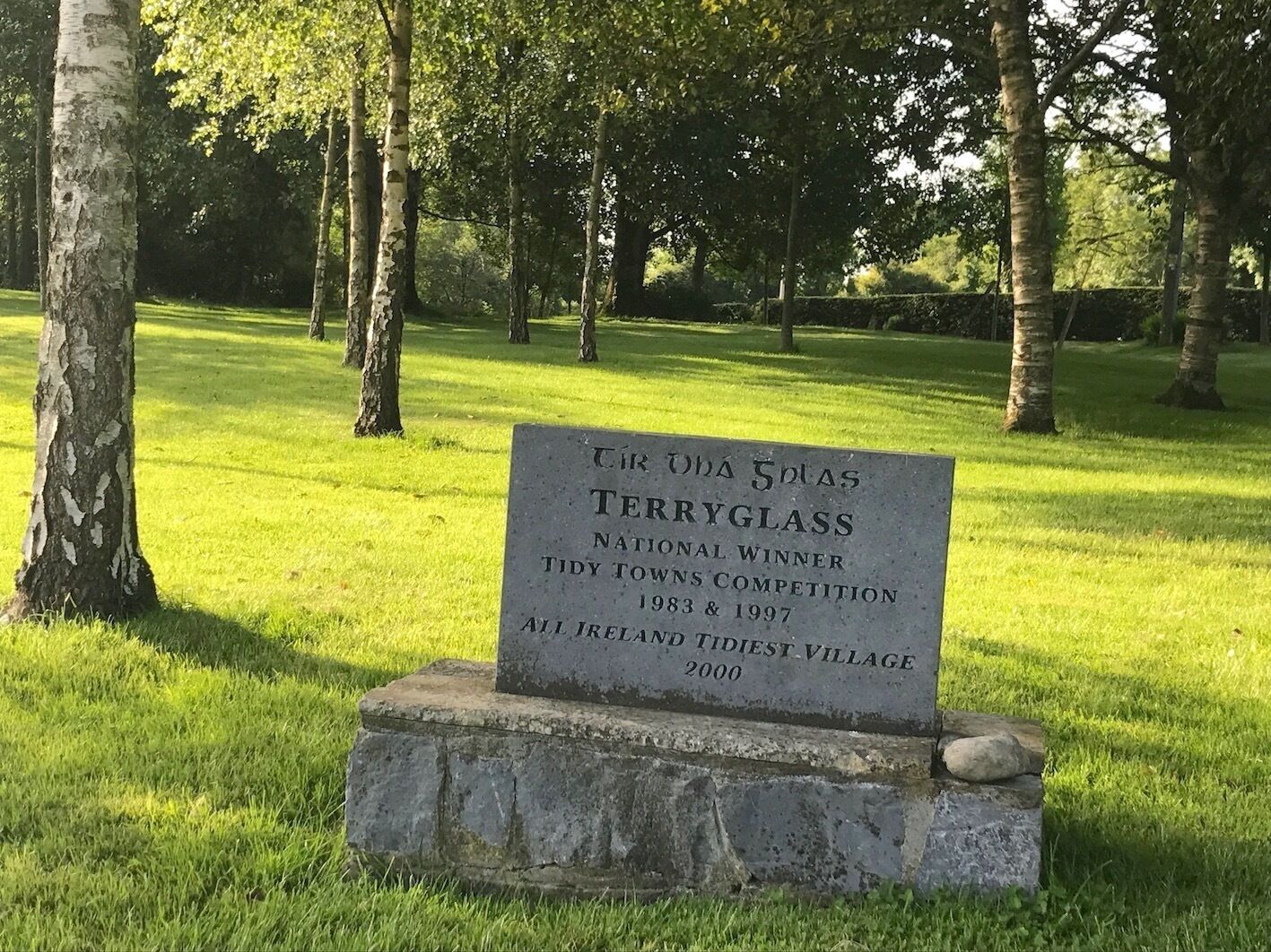 Una placa conmemorativa para Terry Glass, ganador nacional del Concurso de Pueblos Bonitos en 1983 y 1997, ubicada en un parque con césped y árboles.