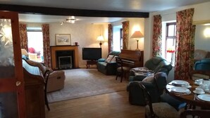 A cozy living room with a fireplace, a television, a piano, and a dining area with a table set for two. - Ashside Cottage,  Royal Hillsborough, Belfast (Lisburn)