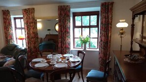 A dining room with a wooden table set for four, surrounded by chairs. There is a window with floral curtains, a lamp on the table, and a piano in the background. - Ashside Cottage,  Royal Hillsborough, Belfast (Lisburn)