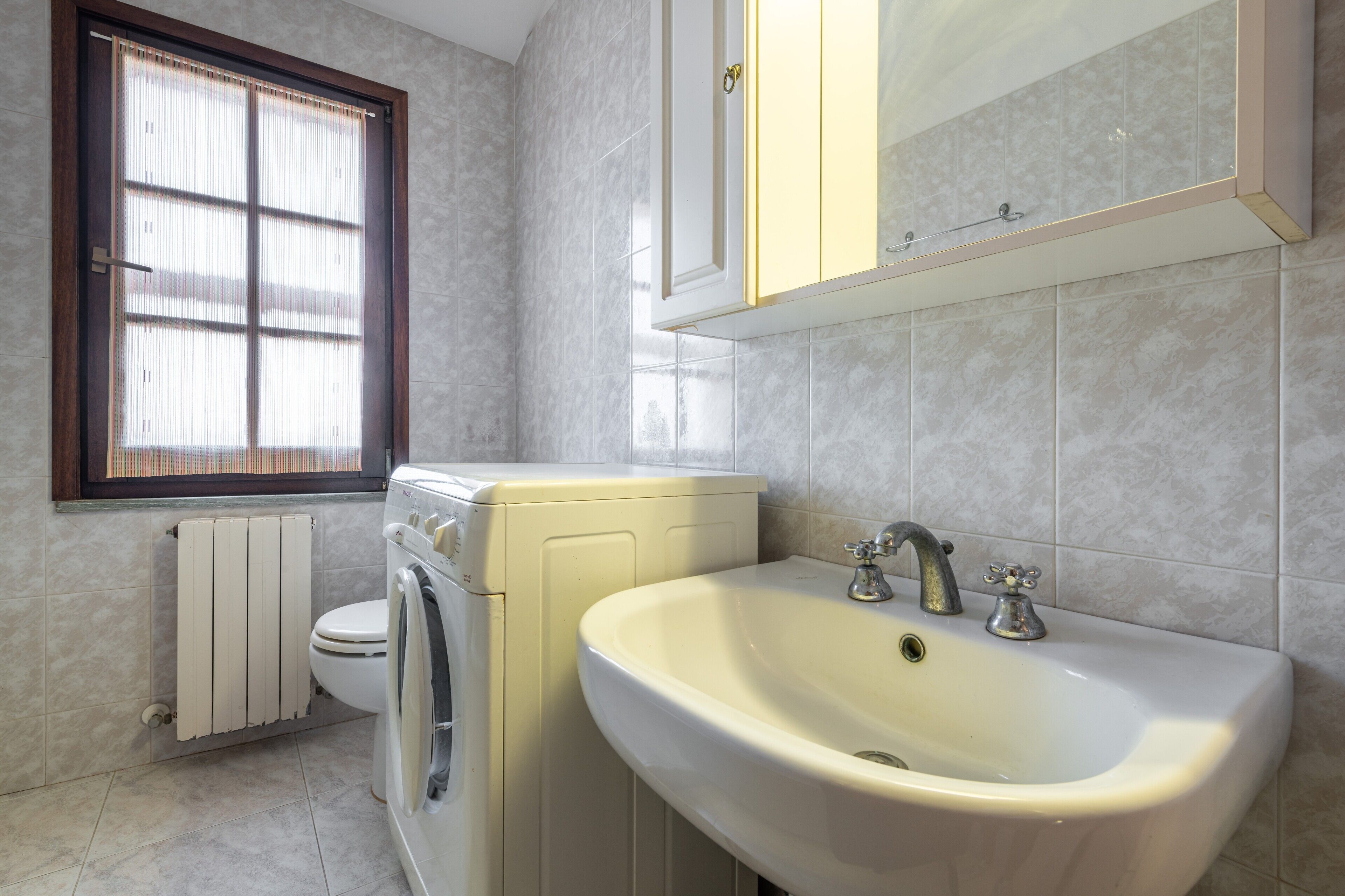A bathroom with a toilet, washing machine, sink, and window with blinds.