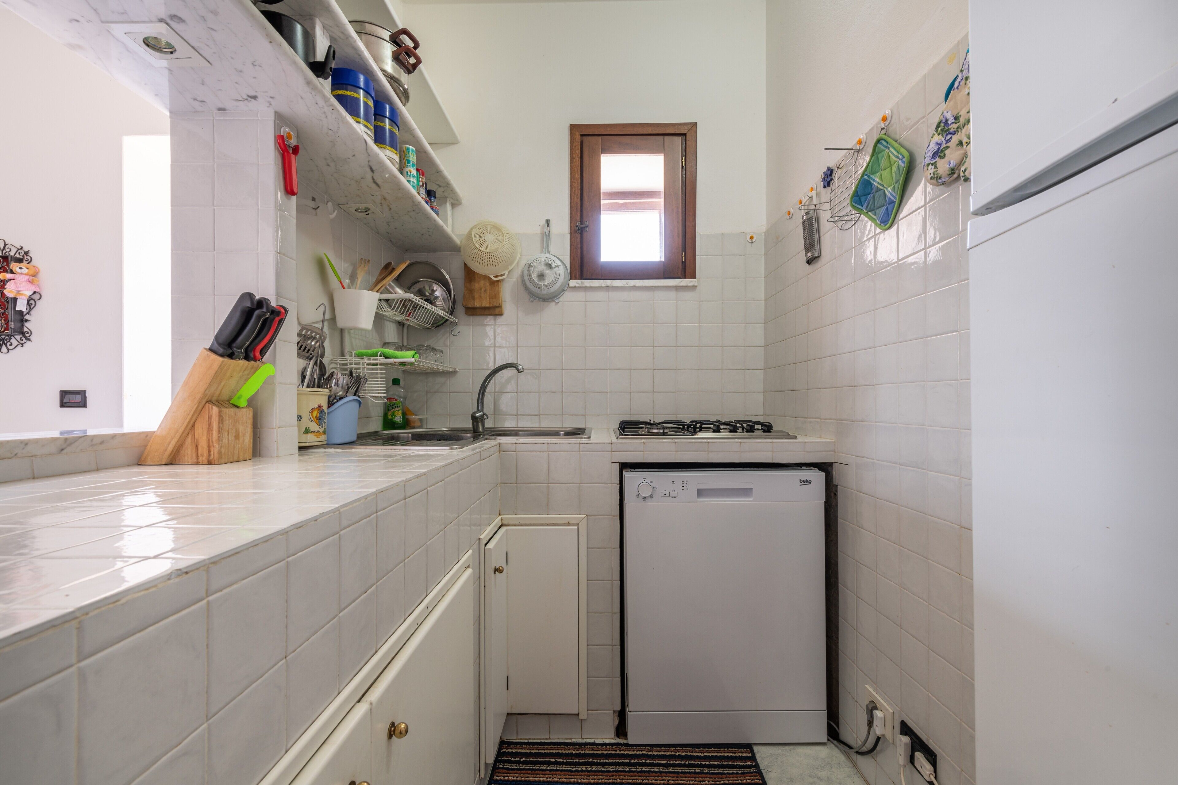 Een kleine keuken met witte tegels, een vaatwasser en een raam.
