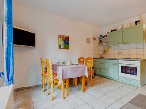 A dining area with a table set for four, wooden chairs, a television mounted on the wall, a painting above the table, and a kitchen with green cabinets and a stove.