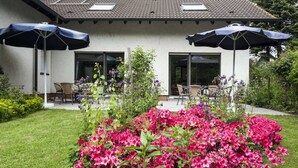 Ein Haus mit einer Terrasse, ausgestattet mit zwei Sonnenschirmen, Sitzgelegenheiten im Freien und einem Garten mit rosa Blumen.