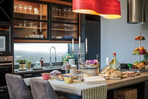 A kitchen with a dining table set for breakfast, featuring bread, pastries, and a fruit display.