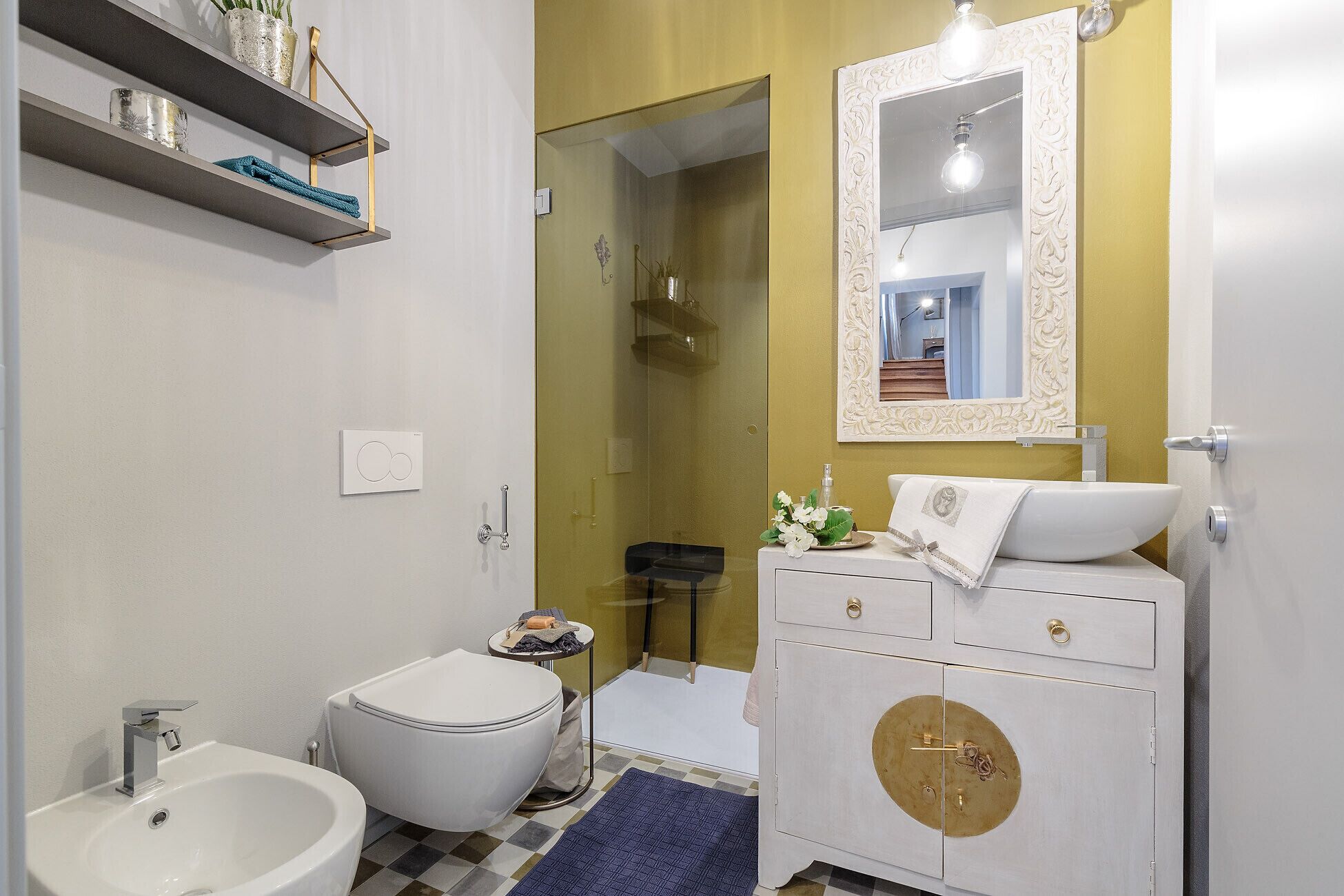 Et moderne badeværelse med toilet, bidet og vask i hvid. Et spejl med detaljeret ramme, et lille bord med toiletartikler, og et skakternet gulv.