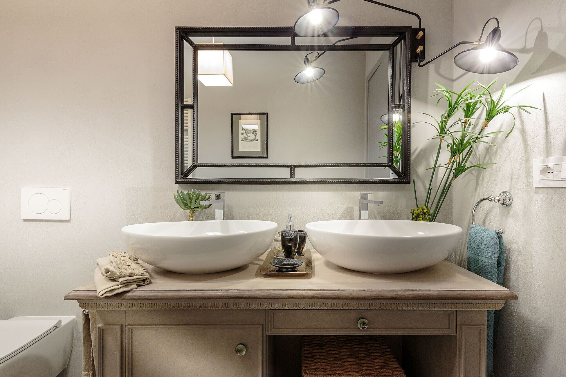 A bathroom with two large white sinks, a mirror with three lights, and a towel rack with a towel.