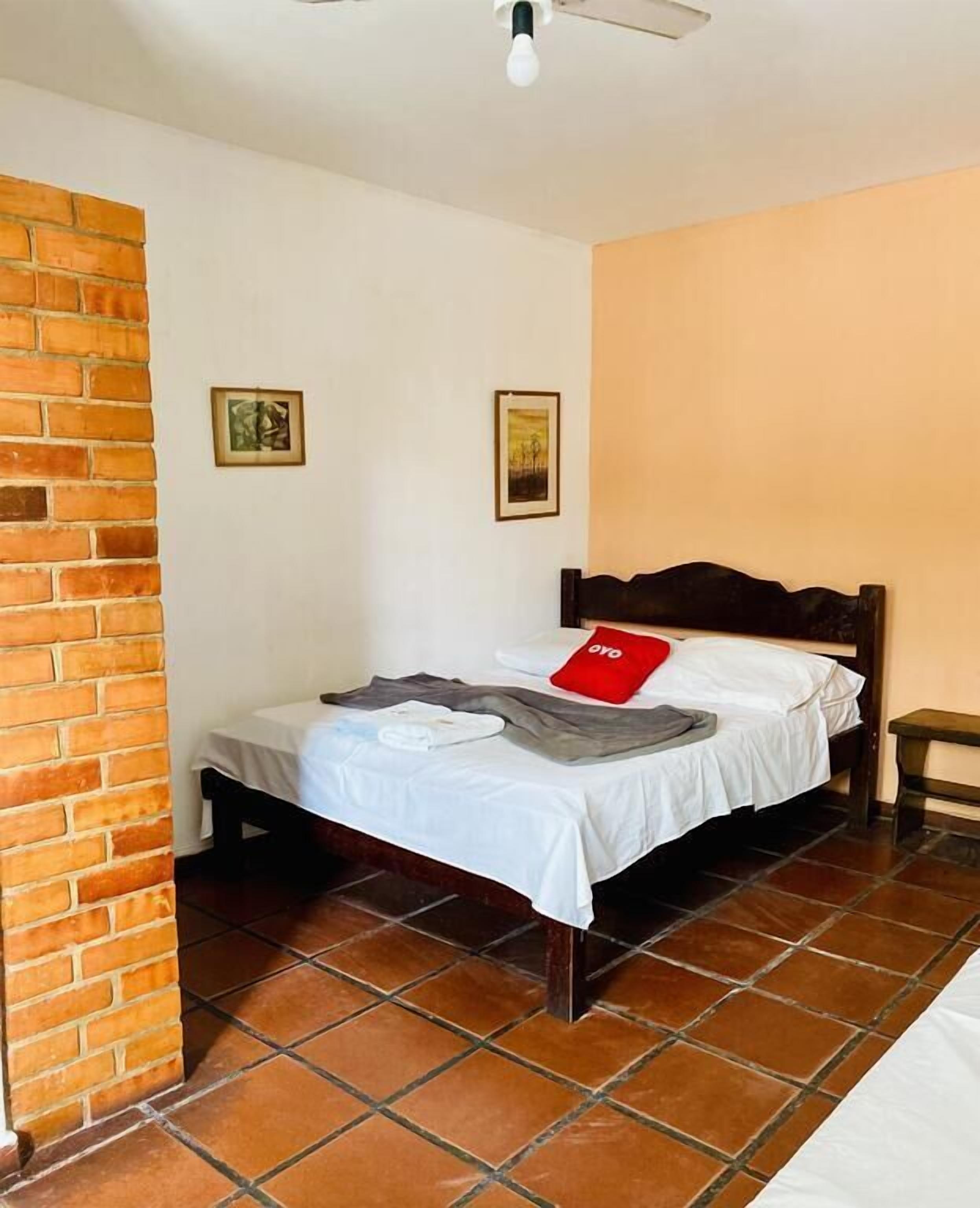 Una cama individual con cabecera de madera, ropa de cama blanca y una almohada roja en una habitación con paredes blancas y un muro de acento de ladrillo.