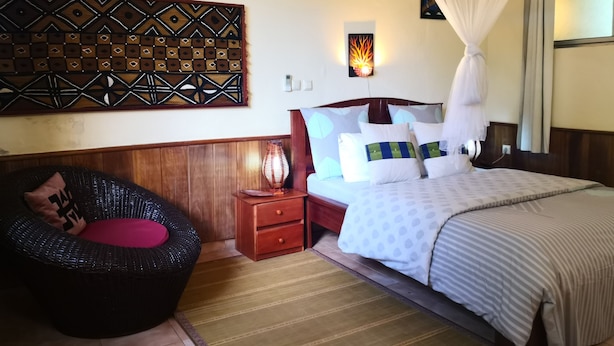 Une chambre d’hôtel comprenant un lit, une table de chevet, un panneau décoratif mural, une lampe fixée au mur, une moustiquaire et un fauteuil en osier.