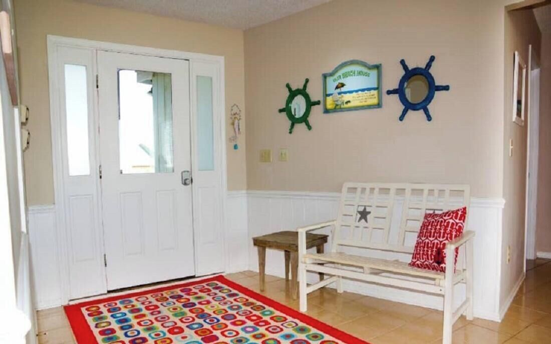 Une pièce avec une porte blanche, un banc blanc orné d’un coussin rouge, un tapis coloré et deux éléments de décoration murale (un vert et un bleu).