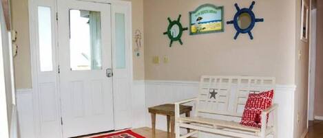 Une pièce avec une porte blanche, un banc blanc orné d’un coussin rouge, un tapis coloré et deux éléments de décoration murale (un vert et un bleu).