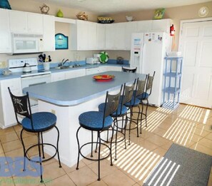 Una cocina con una isla central, armarios blancos, un microondas y un refrigerador. Hay cuatro taburetes azules alrededor de la isla.