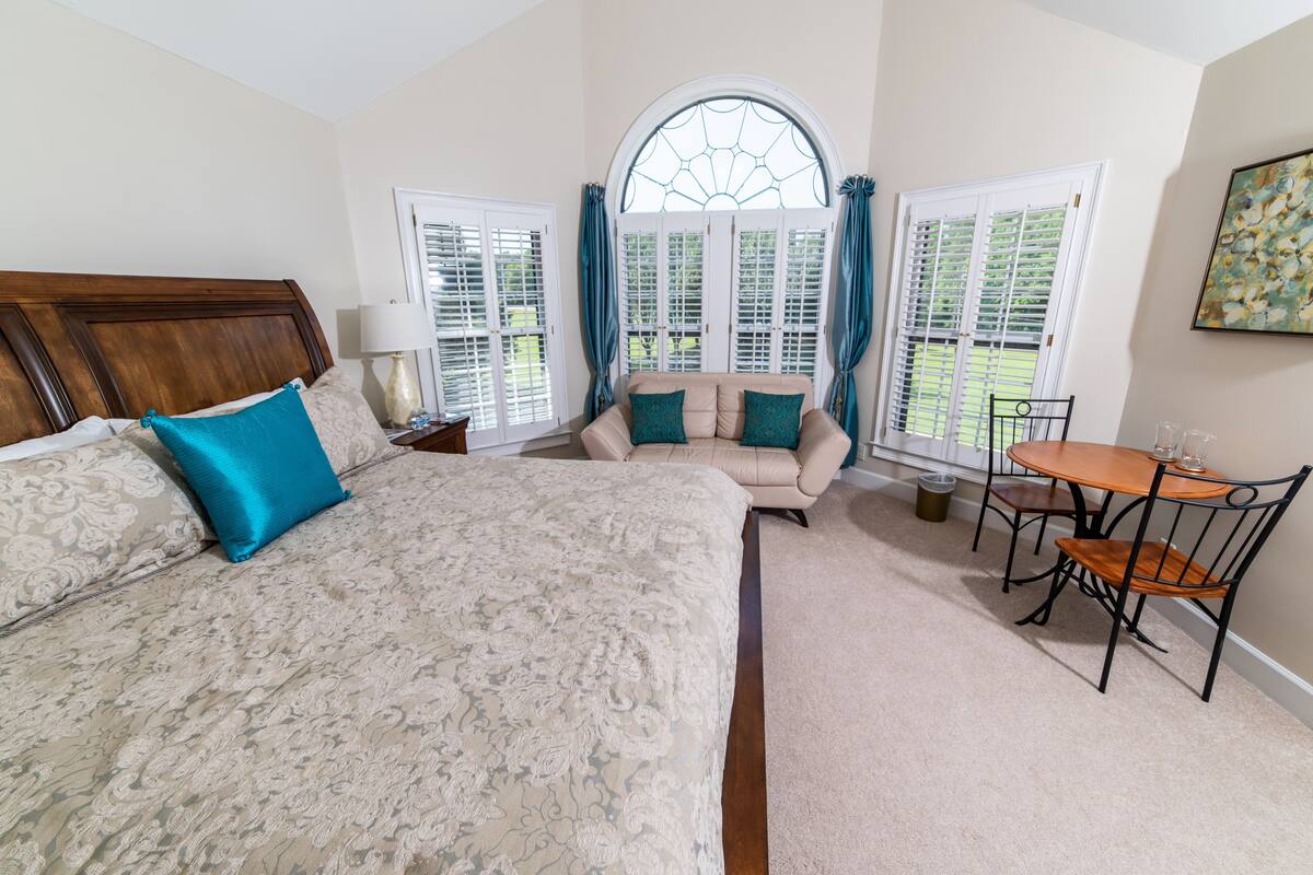 A bedroom with a large bed, a sofa, a dining table with chairs, and a window with shutters.