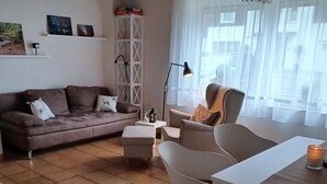 A living room with a brown leather sofa, a white coffee table, and a grey armchair. There is a white shelf with decorative items and a window with sheer curtains. - Central vacation apartment, barrier-free access, please contact us (Braunlage)
