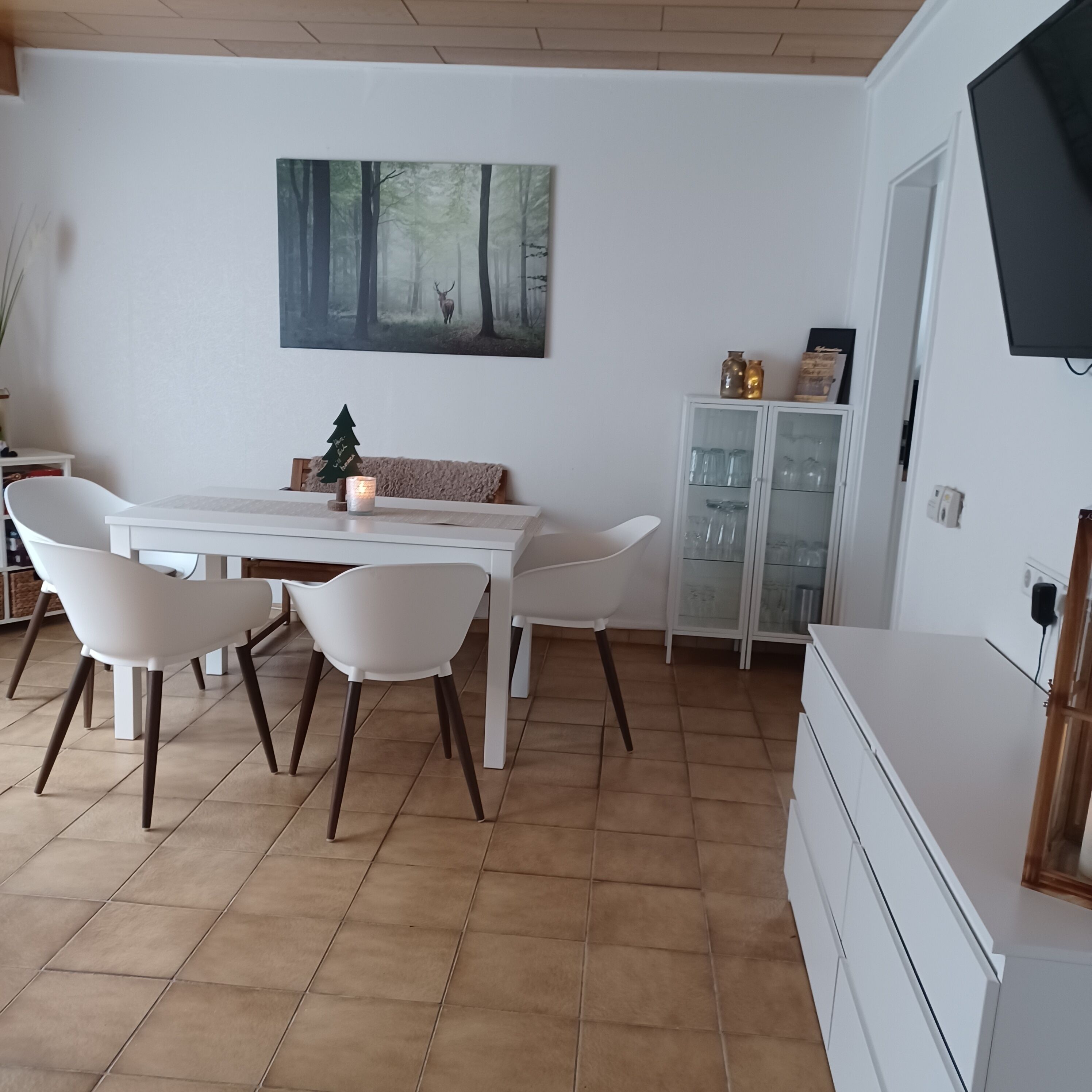 Ein Essbereich mit einem weißen Tisch und Stühlen, einem Bild einer Waldszene, einer kleinen Weihnachtstanne und einem Schrank mit Glastüren.