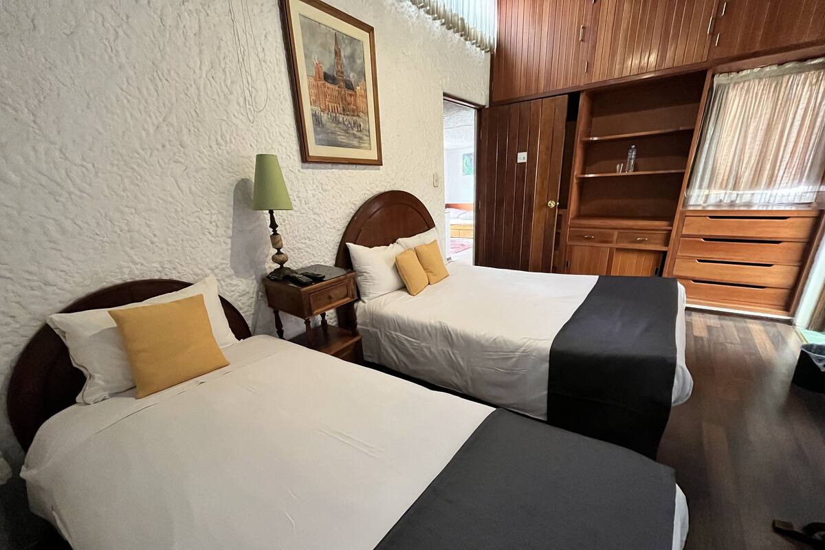 Habitación de hotel con dos camas, un armario de madera, una mesita de noche con lámpara y un cuadro en la pared.