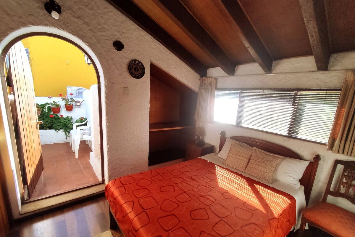 Un dormitorio con cama, mesita de noche, ventana con persianas y vistas a un patio con sillas y plantas.