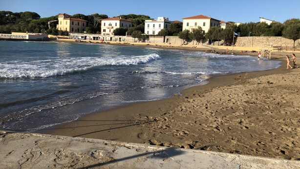 A beach with a sandy shore, waves, and buildings in the background. - Cottage  (Orciano pisano)