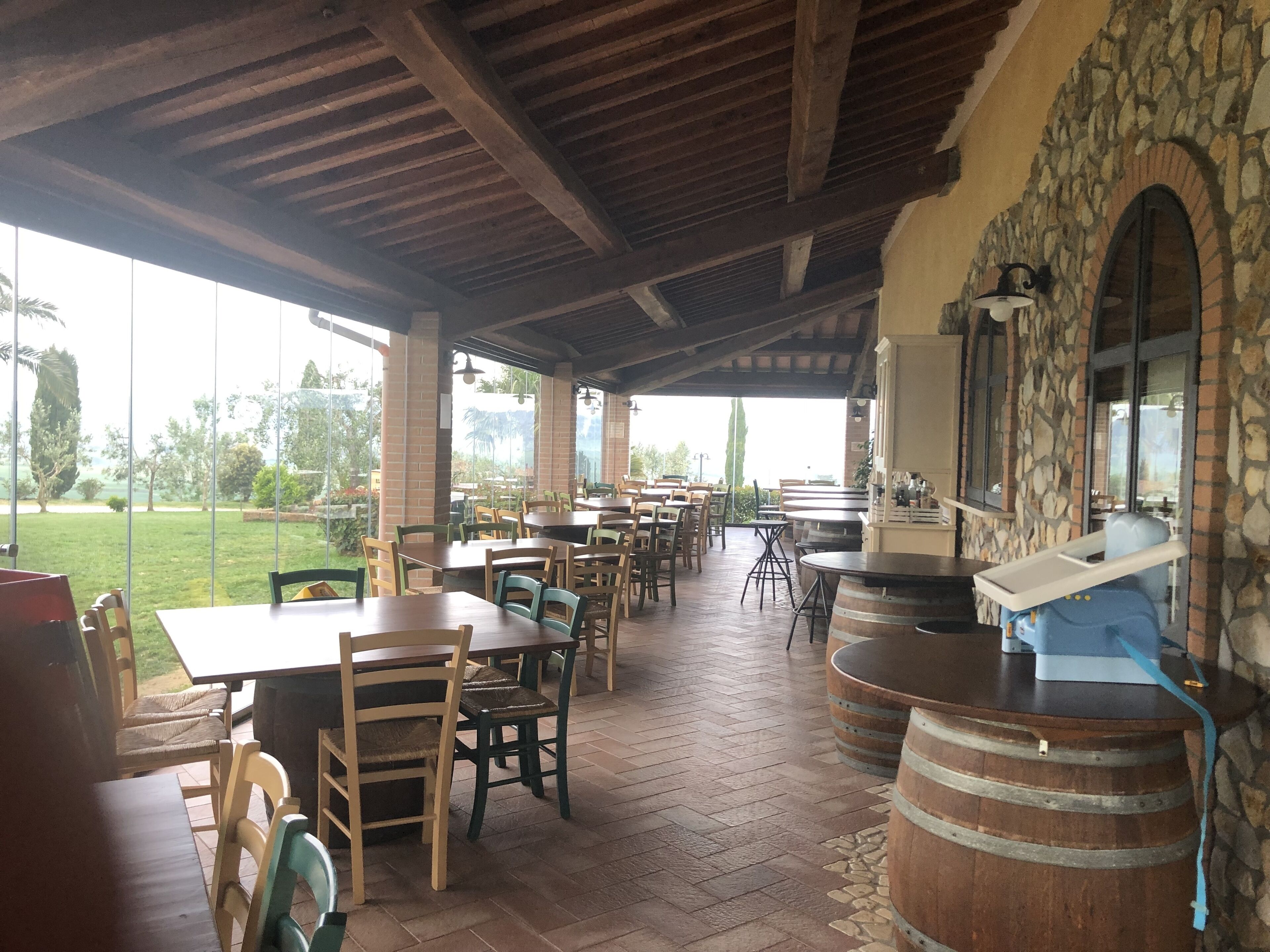 A covered outdoor dining area with brick flooring, wooden tables and chairs, stone walls, and large windows overlooking a garden.