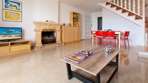 Un vaste espace de vie doté d’une cheminée, d’une table à manger avec des chaises rouges, d’un escalier et d’un téléviseur posé sur un meuble en bois.