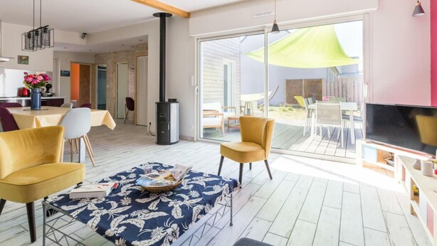 Ein modernes Wohnzimmer mit einer Couch, Sesseln, einem Fernseher und einer großen Schiebeglastür, die auf eine Terrasse im Freien führt.