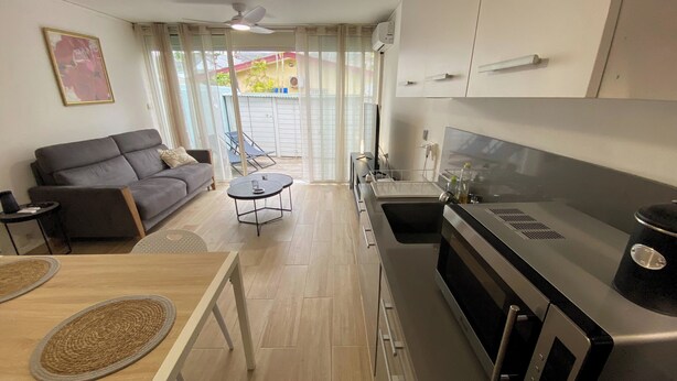 Ein kompakter Wohnraum mit Küchenzeile, einer Couch, einem kleinen Esstisch und einem Balkon mit Blick auf ein Gebäude.
