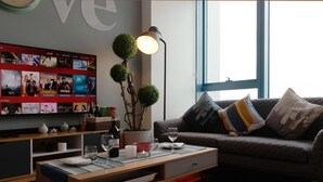 A modern living room with a flat-screen TV displaying a movie selection, a coffee table with glasses and a candle, a sofa with patterned cushions, and a large window providing natural light. - Posh Entrata SOHO Suite-- Festival Mall view (Alabang, Muntinlupa City)