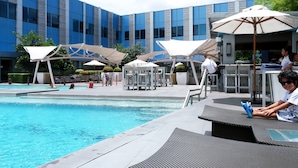 A pool area with lounge chairs and umbrellas, a building with blue and grey facade, and a clear sky. - Posh Entrata SOHO Suite-- Festival Mall view (Alabang, Muntinlupa City)