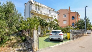 A residential street with a white car parked in front of a building with a balcony. - Apartments Karleusa (Rogoznica)
