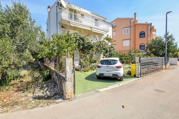 A residential street with a white car parked in front of a building with a balcony. - Apartments Karleusa (Rogoznica)