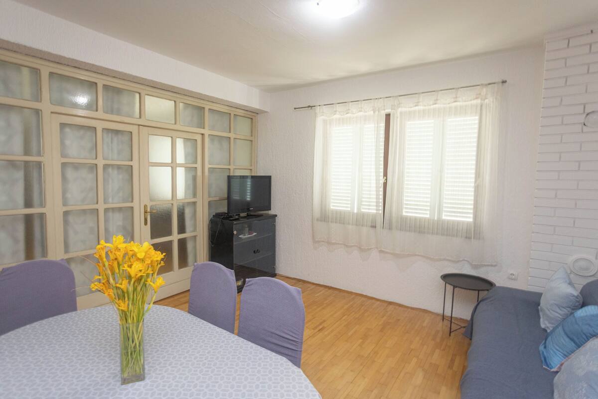 A room with a dining table, chairs, a sofa, a television on a cabinet, and a window with curtains.