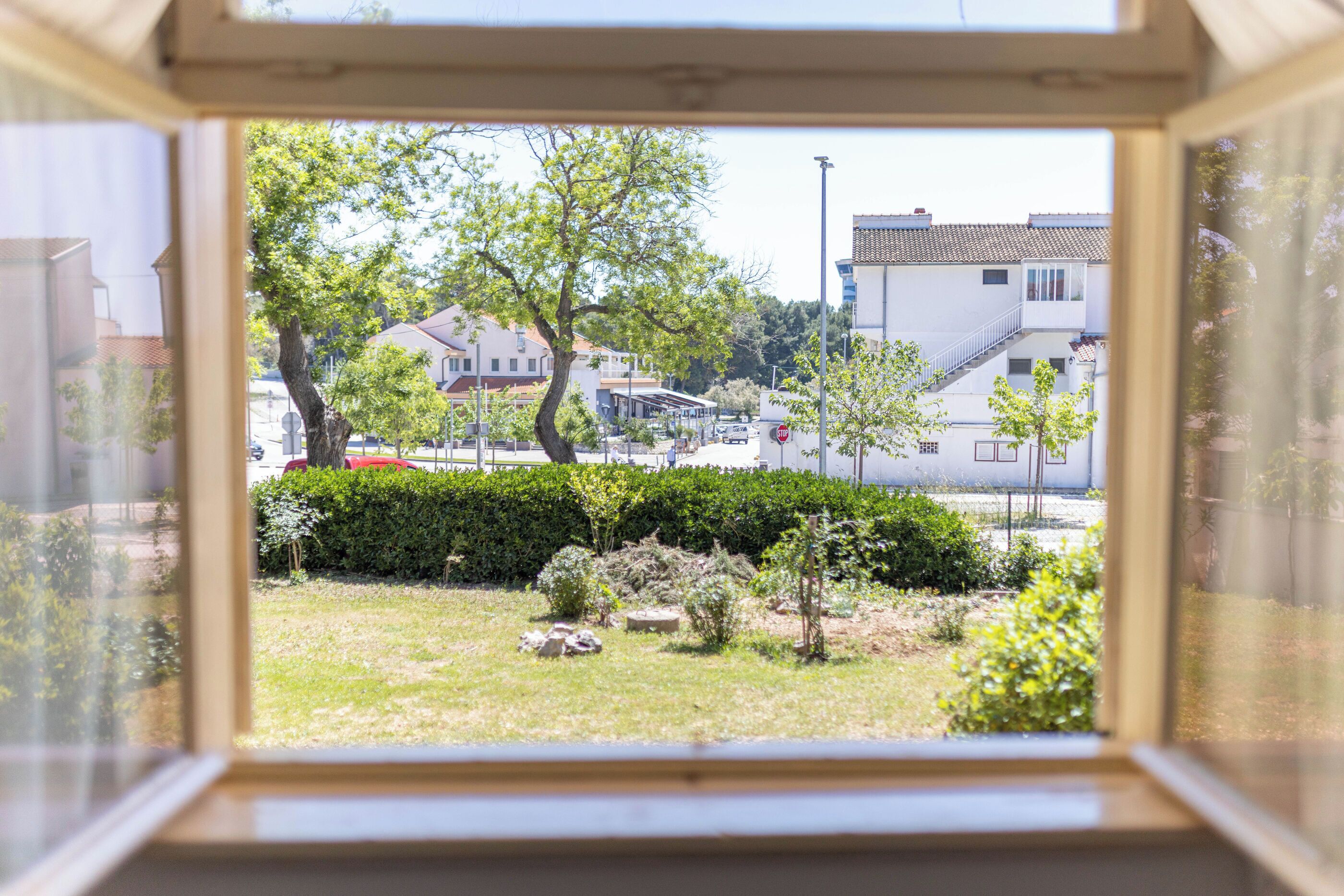 A view through a window of a residential area with houses, a street, and a grassy area with bushes and trees.