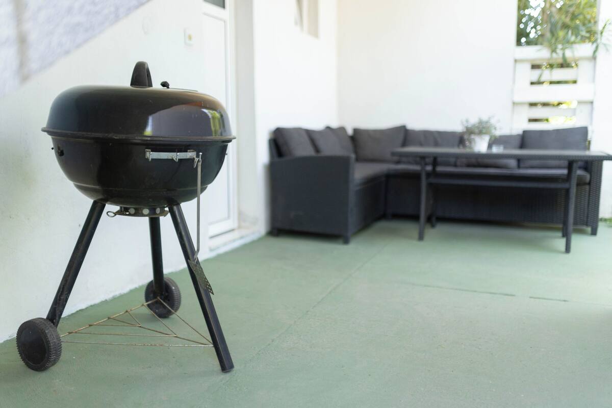A black barbecue grill on a stand with wheels, positioned on a green outdoor floor. In the background, there is a dark-colored sofa and a table with a potted plant.