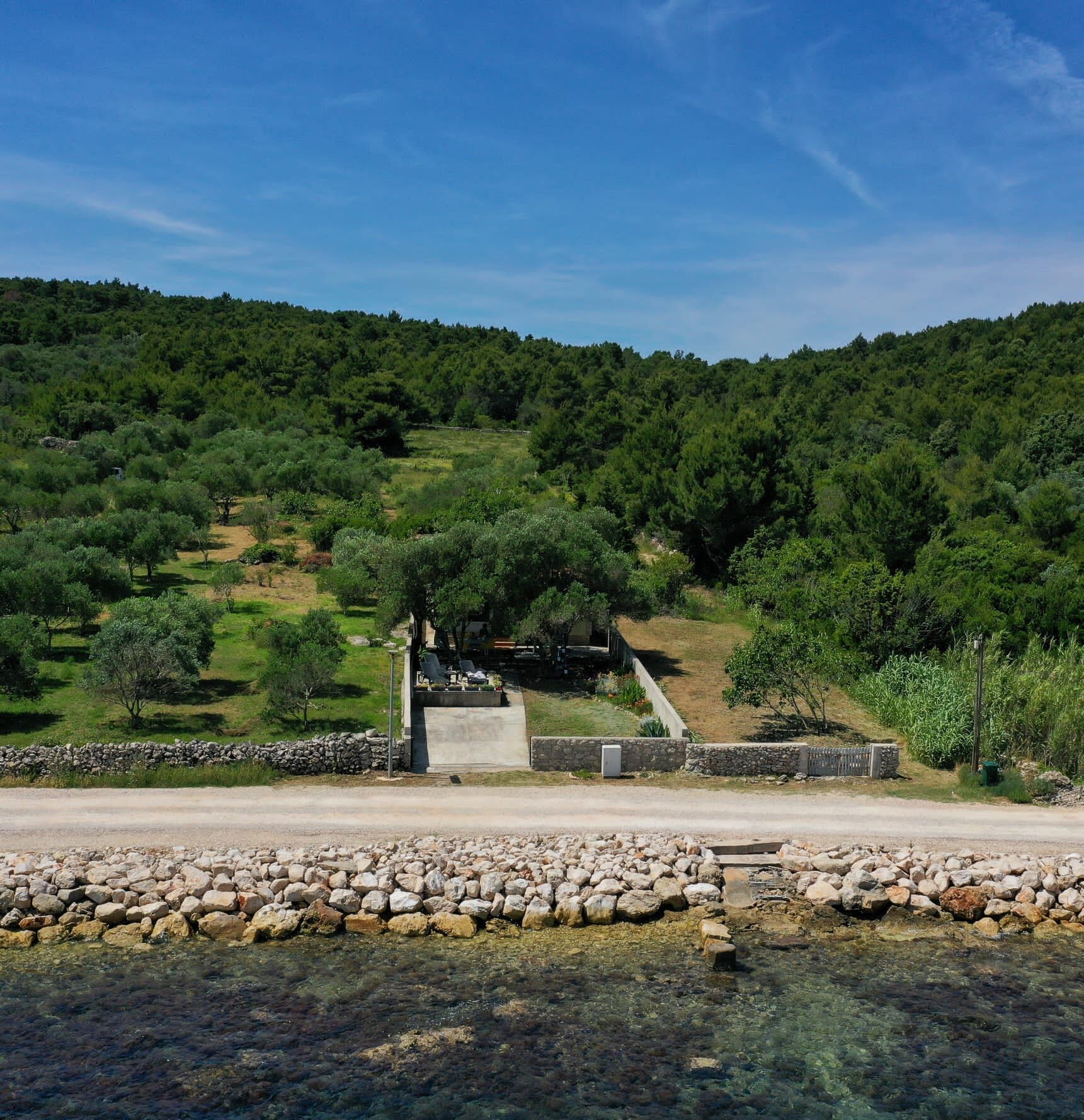 A rural landscape with a stone wall, a small building with a paved area, and a clear view of a forested hill.