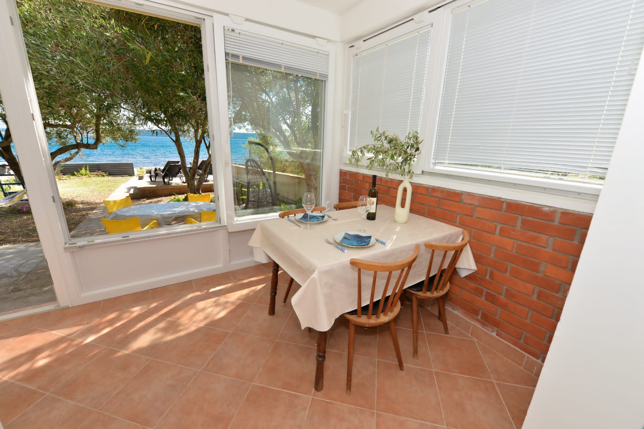 A dining area with a table set for four, large windows with blinds, a brick wall, and a view of the sea.