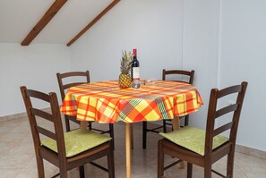 Ein runder Tisch mit kariertem Tischtuch, vier Holzstühle mit grünen Kissen, eine Weinflasche und eine Ananas.