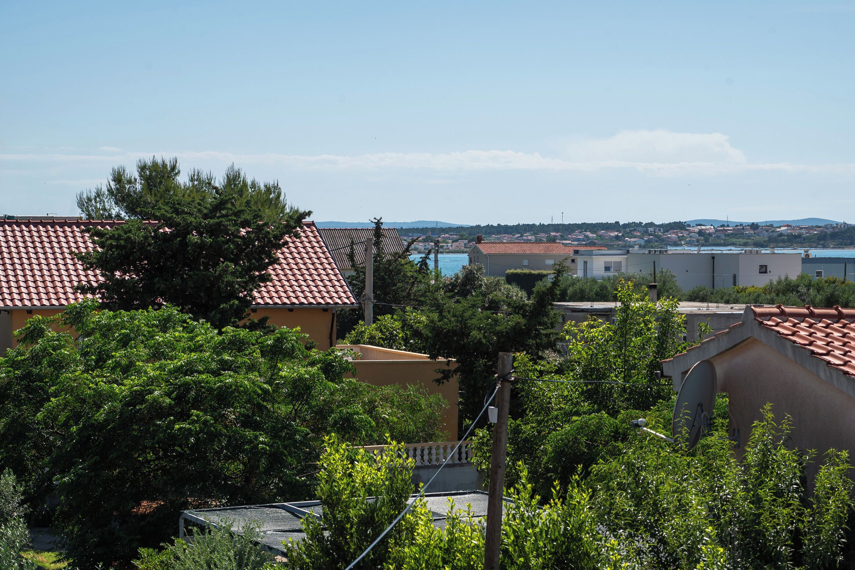 A residential area with terracotta-roofed houses, lush greenery, and a distant view of a body of water and hills.