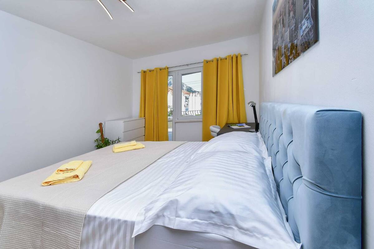 A hotel room with a bed, a window with yellow curtains, a small plant, and a painting on the wall.