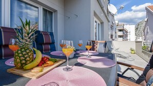 Un balcón con mobiliario exterior, una mesa con fruta y bebidas, y vista panorámica de la ciudad.