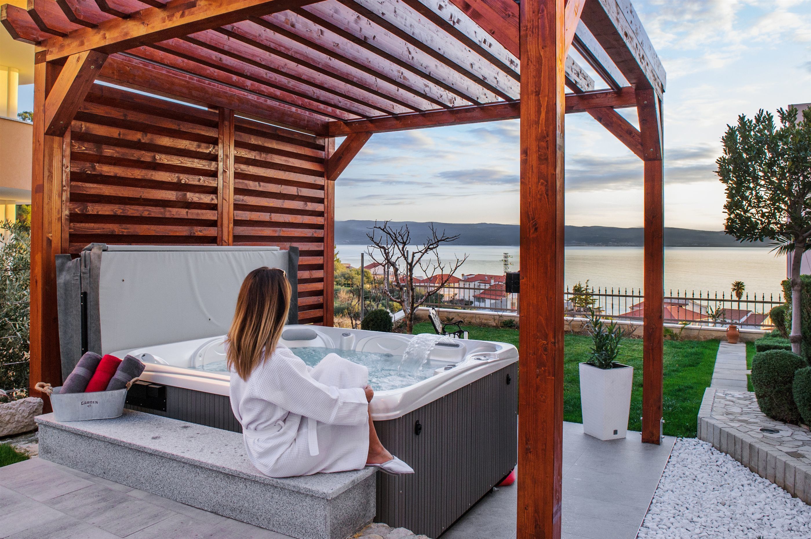 A person relaxing in a hot tub under a wooden pergola with a view of a lake and buildings in the background.