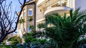 A modern multi-story building with balconies, surrounded by lush greenery and a tree. - Apartment and Rooms Ana (Podstrana)