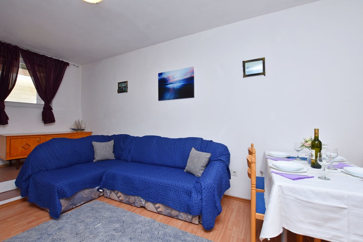 A living room with a blue sectional sofa, a dining table set for two, and a small cabinet.