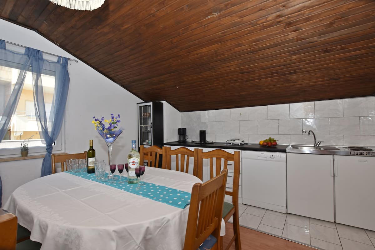A dining area with a table set for two, wooden chairs, a kitchen with white cabinets, and a wooden ceiling.