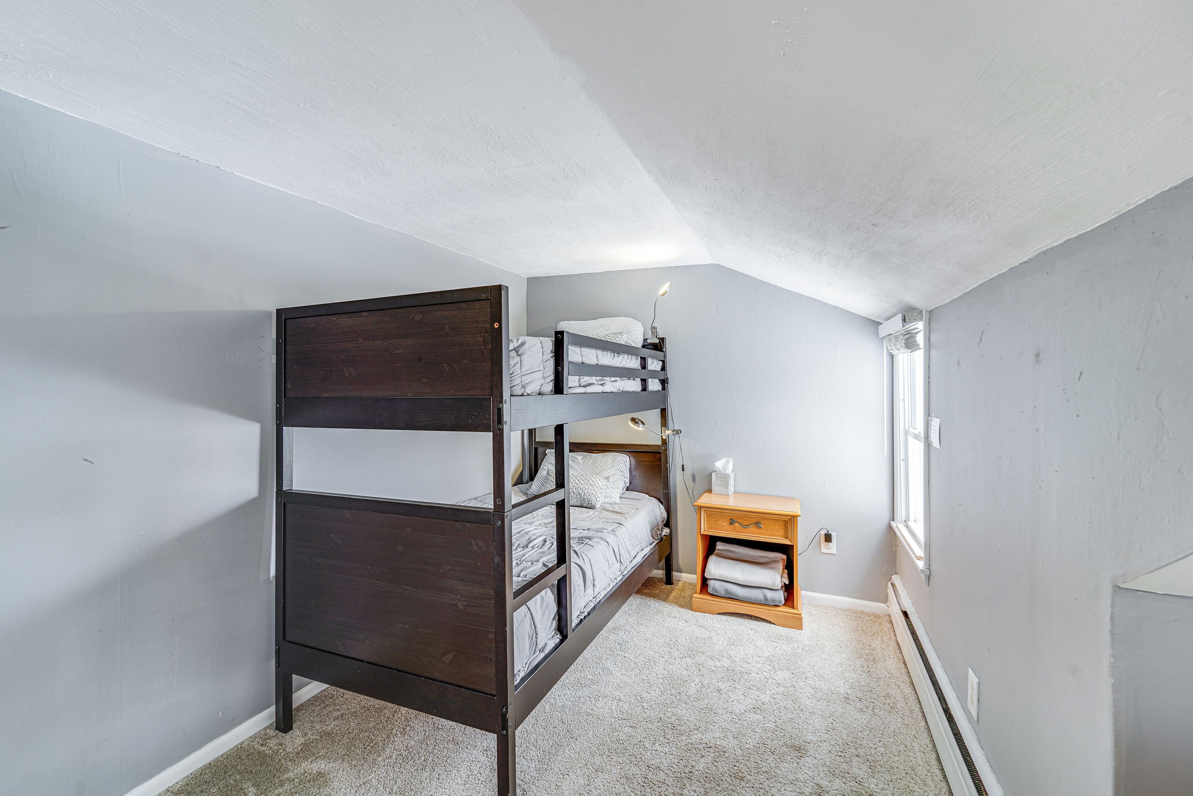 Une chambre avec un lit superposé, une petite table de chevet en bois, une lampe et une fenêtre.