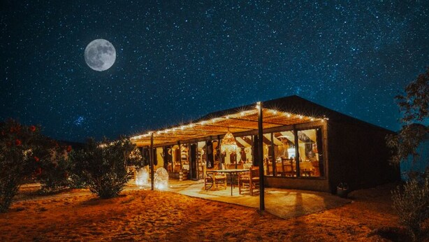 Un edificio con un portico coperto, un interno visibile con tavoli e sedie, e una grande luna nel cielo notturno.